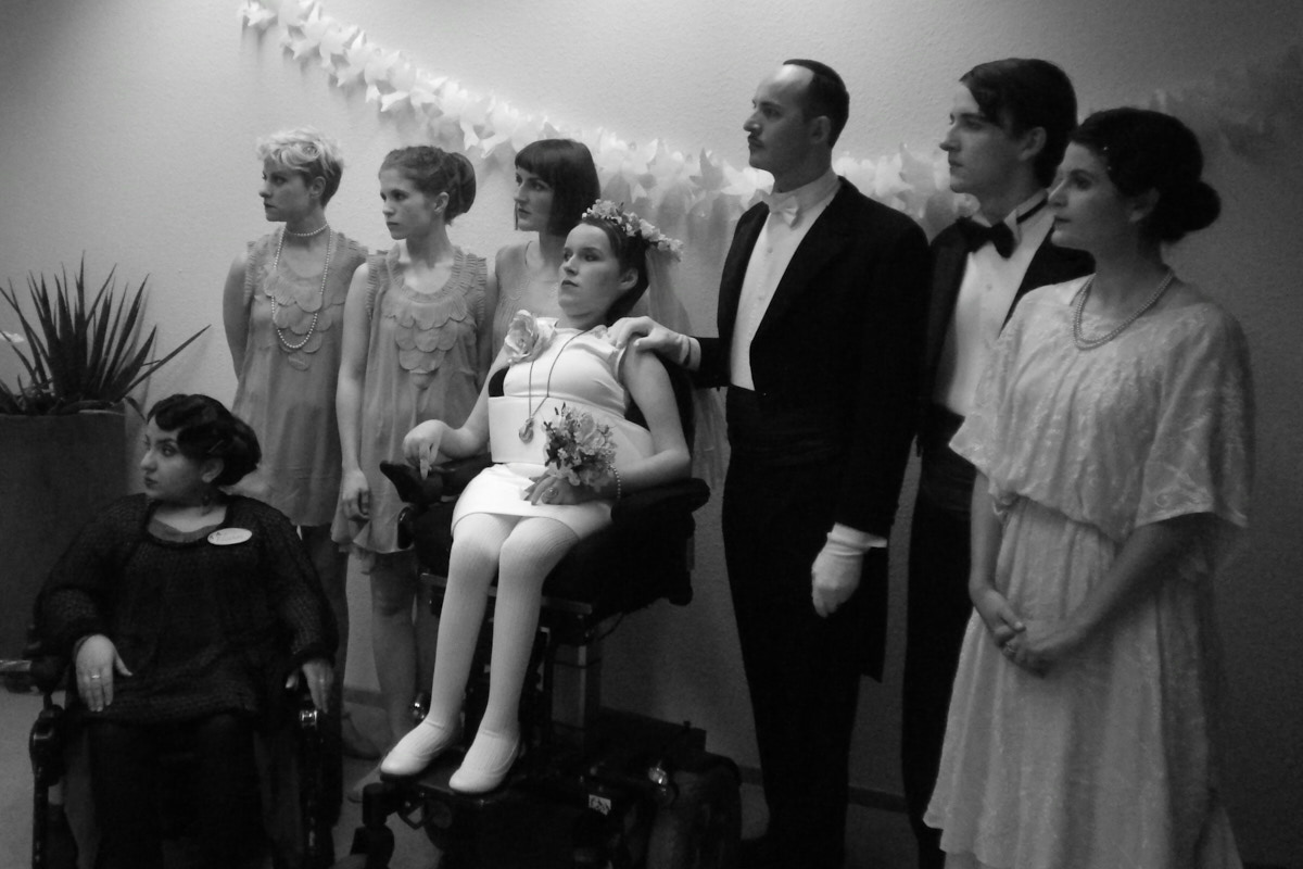 8 Menschen in feierlicher Kleidung stehen in einer Reihe. Eine Frau trägt ein weisses Brautkleid. Sie sitzt im Rollstuhl. Ein Mann trägt einen schwarzen Frack. Foto: dorisdean