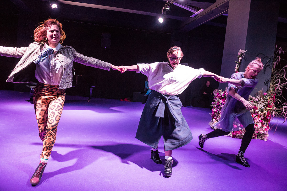 3 Schauspielerinnen halten sich an den Händen und laufen durch einen violett beleuchteten Raum, Foto: Christian Knieps