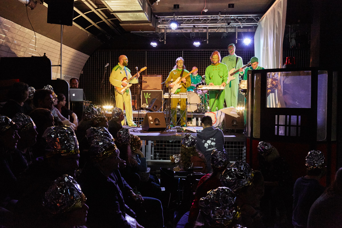 Eine Gruppe Menschen in grünen Kostümen spielen Musik auf einer Bühne. Die Zuschauer davor tragen Hüte aus silberne Folie. Foto: Christian Martin