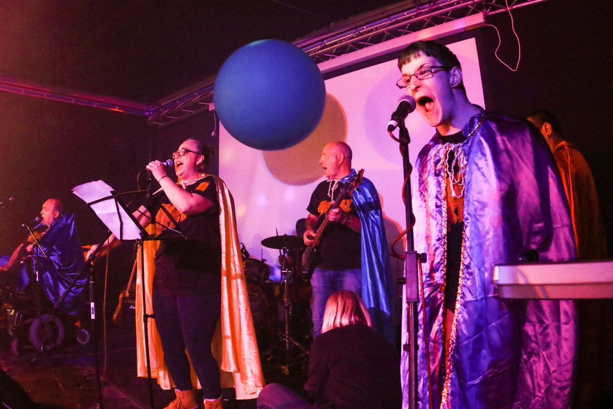 4 Musiker mit bunten Umhängen singen auf einer Bühne. Oben hängt ein blauer Ball. Foto: Jonathan Dunn
