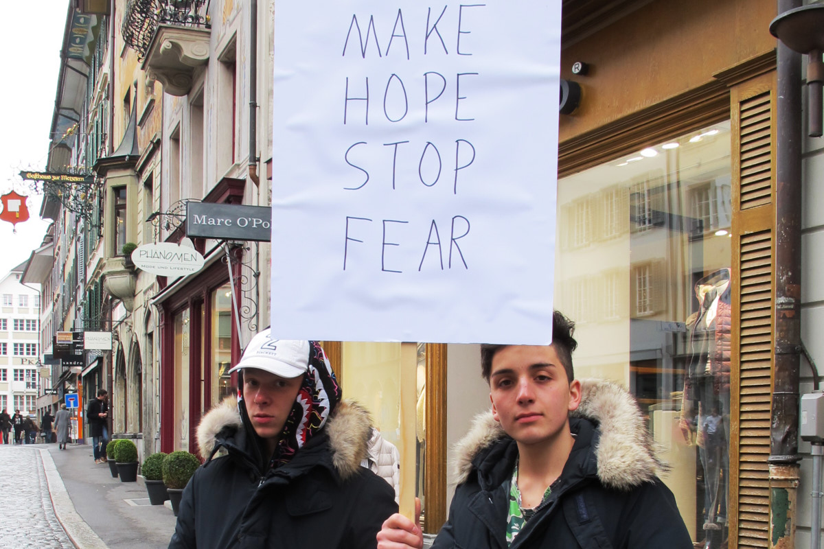 Zwei junge Menschen in einer Straße. Einer hält ein Schild hoch, darauf steht in Englisch "Macht Hoffnung. Stoppt Angst." Foto: the vacuumcleaner